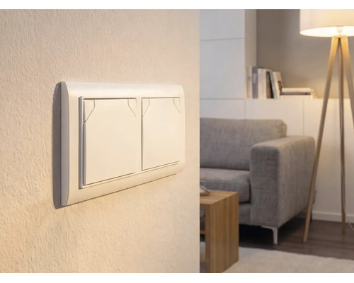 Dubbele lichtschakelaar op een muur in een lichte woonkamer met bank en vloerlamp.