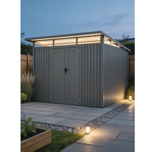 Modern metalen tuinhuis met lessenaarsdak, bovenlichten en dubbele deur op een bestraat oppervlak in een helder verlichte tuin.