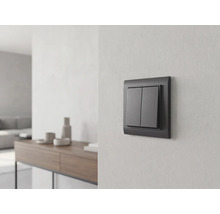 Antracietkleurige serieschakelaar aan een lichte wand in een lichte, modern ingerichte woonkamer met dressoir op de achtergrond.