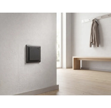 Donkergrijze moderne lichtschakelaar op een lichte muur in een minimalistische, lichte gang met houten vloer.