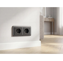 Antracietkleurige dubbele wandcontactdoos op witte muur boven plint in lichte woonkamer met houten vloer.