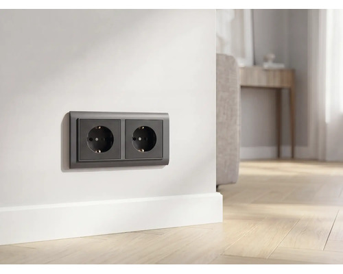 Antracietkleurige dubbele wandcontactdoos op witte muur boven plint in lichte woonkamer met houten vloer.