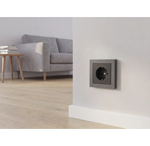 Antraciet stopcontact op een witte muur in een lichte woonkamer met een grijze bank en houten vloer.