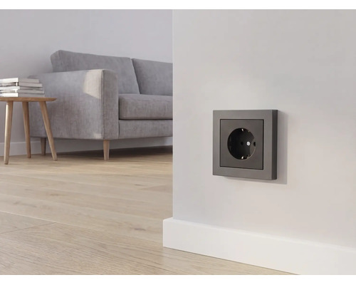Antraciet stopcontact op een witte muur in een lichte woonkamer met een grijze bank en houten vloer.