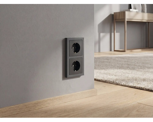 Dubbele contactdoos in antraciet op een grijze muur in een modern ingerichte kamer met houten vloer.