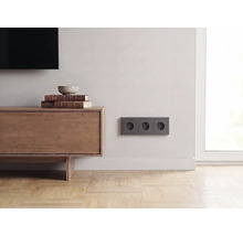 Drievoudig stopcontact in antraciet aan een lichte wand in een modern ingerichte woonkamer met houten dressoir en parketvloer.