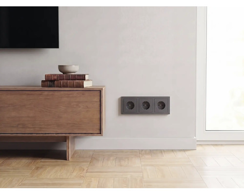 Drievoudig stopcontact in antraciet aan een lichte wand in een modern ingerichte woonkamer met houten dressoir en parketvloer.