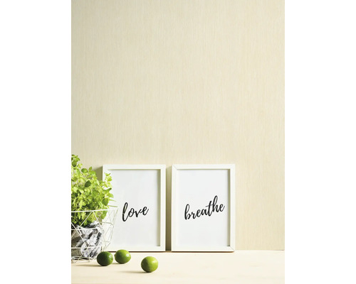 Lichtbeige behang met een fijne verticale structuur achter witte fotolijstjes en een groene plant.