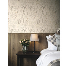 Vliesbehang met bloemenpatroon in een slaapkamer boven een houten wandpaneel met nachtkastje, lamp en bloemen.