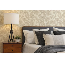 Slaapkamer met vliesbehang met palmbladpatroon, houten nachtkastje met witte tafellamp en bed met verschillende kussens en gebreide deken.