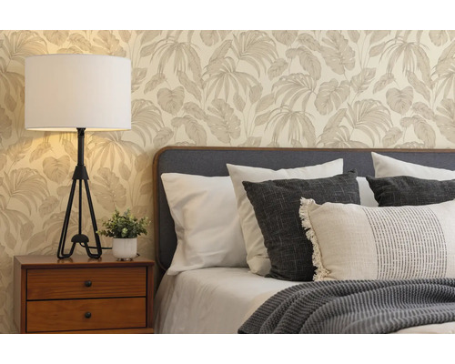 Slaapkamer met vliesbehang met palmbladpatroon, houten nachtkastje met witte tafellamp en bed met verschillende kussens en gebreide deken.