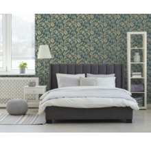 Vliesbehang met goudkleurig palmbladpatroon achter een grijs gestoffeerd bed in een moderne, lichte slaapkamer.