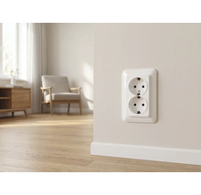 Dubbele wandcontactdoos met randaarde op een lichte muur in een moderne woonkamer met houten vloer en fauteuil.