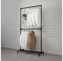 Zwart kledingrek van industriële buizen met twee stangen, met witte overhemden, een beige jas en een hangplant tegen een witte bakstenen muur.