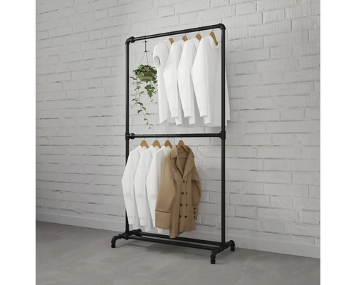 Zwart kledingrek van industriële buizen met twee stangen, met witte overhemden, een beige jas en een hangplant tegen een witte bakstenen muur.