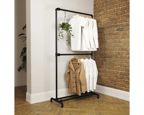 Tweelaags kledingrek van zwarte metalen buizen in industriële stijl voor een bakstenen muur, met witte kleding en een hangplant.