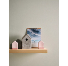 Gestructureerd vliesbehang in lichtgroen op een muur achter een houten wandplank met decoratieartikelen zoals kleine huisjes en een klembord.