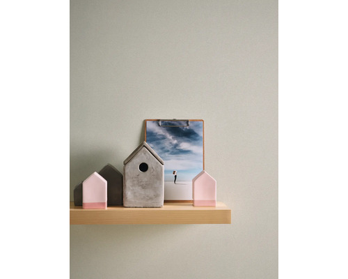 Gestructureerd vliesbehang in lichtgroen op een muur achter een houten wandplank met decoratieartikelen zoals kleine huisjes en een klembord.
