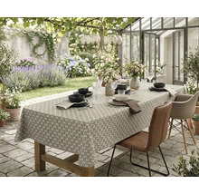Gedekte houten tuintafel met een tafelkleed met patroon op een stenen terras voor een plantenkas, omringd door tuinstoelen.