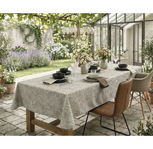 Gedekte houten tafel op een stenen terras met een tafelkleed met bladmotief, leren stoelen en bloemstukken in een tuin.