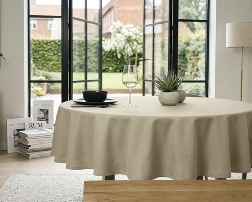 Rond beige tafelkleed op een eettafel in een lichte kamer met uitzicht op een tuin door grote ramen.