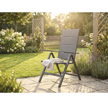 Inklapbare tuinstoel met hoge rugleuning op een stenen terras in een zonnige tuin met bloemen en een houten hek.