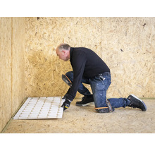 Een man legt witte isolatieplaten op een houten vloer. Hij draagt kniebeschermers en handschoenen in een ruimte met houten wanden.