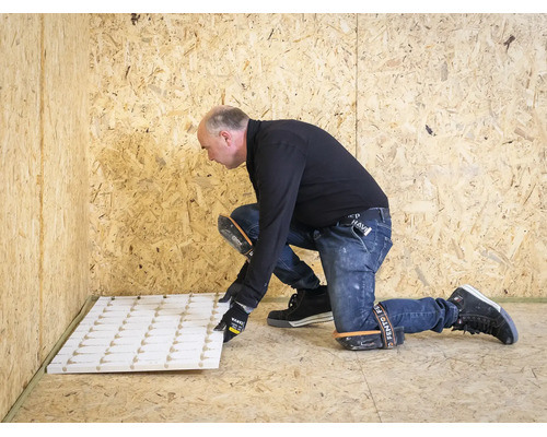 Een man legt witte isolatieplaten op een houten vloer. Hij draagt kniebeschermers en handschoenen in een ruimte met houten wanden.