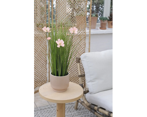 Kunstgras met roze bloemen in een geribbelde pot op een ronde houten tafel naast een fauteuil met wit kussen.