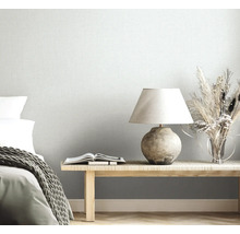 Lichtgrijs vliesbehang met linnenlook in een lichte slaapkamer met houten bank, tafellamp, droogbloemen en bed met gebreide deken.