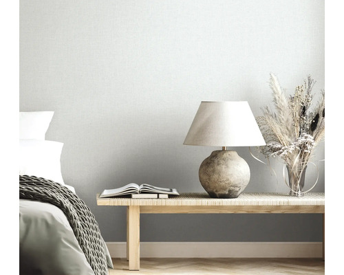 Lichtgrijs vliesbehang met linnenlook in een lichte slaapkamer met houten bank, tafellamp, droogbloemen en bed met gebreide deken.