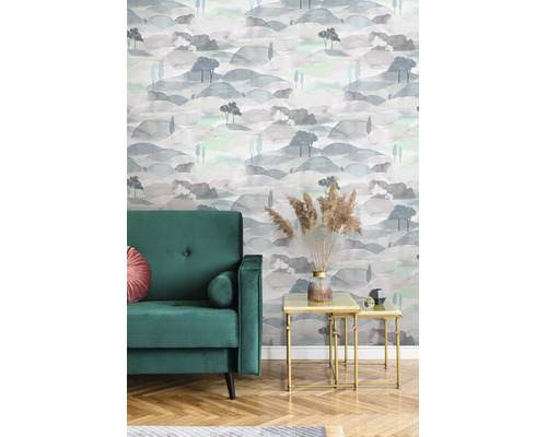 Vliesbehang met landschapspatroon in aquarel-look, heuvels en bomen in een woonkamer met groene fauteuil en bijzettafels.