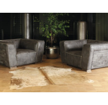 Twee grijze leren fauteuils met siernaden op een parketvloer, een koeienhuid vloerkleed op de voorgrond en een kamerplant in een metalen pot.