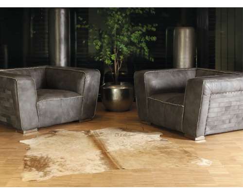 Twee grijze leren fauteuils met siernaden op een parketvloer, een koeienhuid vloerkleed op de voorgrond en een kamerplant in een metalen pot.