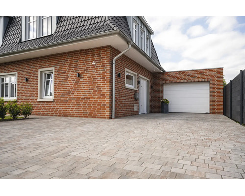 Exterieur van een bakstenen huis met een witte garagedeur en een grote oprit bestraat met rechthoekige stenen.