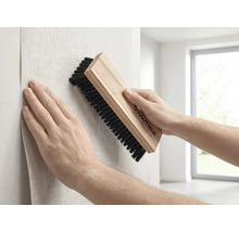 Een persoon strijkt behang glad met een Rautner behangborstel met houten handvat en donkere haren tijdens het behangen van een muur.