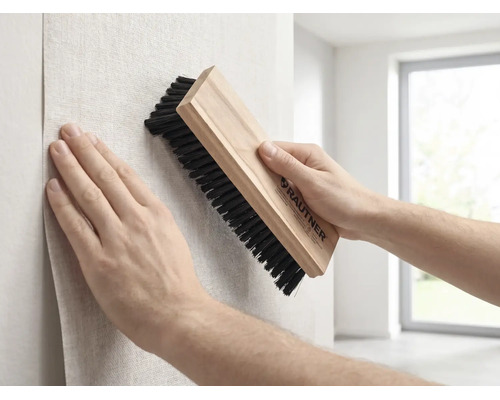Een persoon strijkt behang glad met een Rautner behangborstel met houten handvat en donkere haren tijdens het behangen van een muur.
