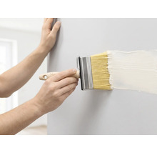 Een persoon schildert een muur met een brede blokkwast en witte verf.