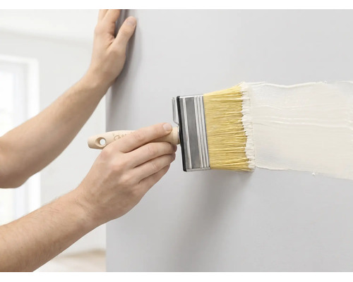 Een persoon schildert een muur met een brede blokkwast en witte verf.
