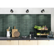 Moderne keuken met donkergroene achterwand van rechthoekige tegels, lichte houten kasten en keukenbenodigdheden op het aanrecht.
