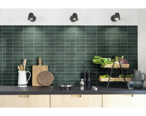 Moderne keuken met donkergroene achterwand van rechthoekige tegels, lichte houten kasten en keukenbenodigdheden op het aanrecht.