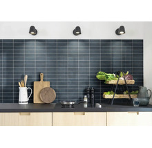 Moderne keuken met een donkere betegelde achterwand, lichte houten kasten en keukengerei onder drie wandspots.