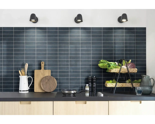 Moderne keuken met een donkere betegelde achterwand, lichte houten kasten en keukengerei onder drie wandspots.