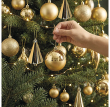 Een hand hangt een glanzende, ronde kerstbal in een versierde kerstboom met verschillende ornamenten en een lichtsnoer.