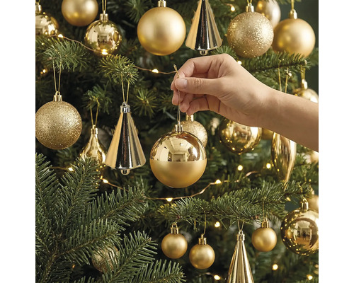 Een hand hangt een glanzende, ronde kerstbal in een versierde kerstboom met verschillende ornamenten en een lichtsnoer.