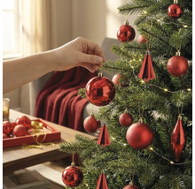 Hand hangt een rode kerstbal in een versierde kerstboom met lichtjes en verschillende rode ornamenten in een lichte kamer.