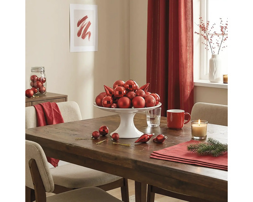 Feestelijk gedekte houten tafel met een witte etagère vol rode kerstballen, een kaars en adventsdecoratie.