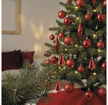 Kerstboom versierd met verschillende rode ornamenten en warmwitte lampjes in een lichte woonkamer.