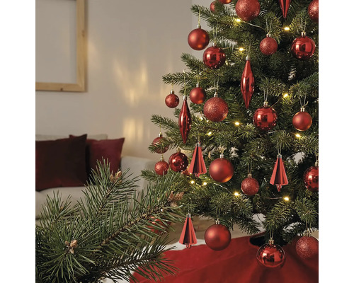 Kerstboom versierd met verschillende rode ornamenten en warmwitte lampjes in een lichte woonkamer.