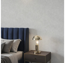 Goudkleurige tafellamp op een nachtkastje in een slaapkamer met een blauw gestoffeerd bed en grijze kussens voor een muur met textuur.